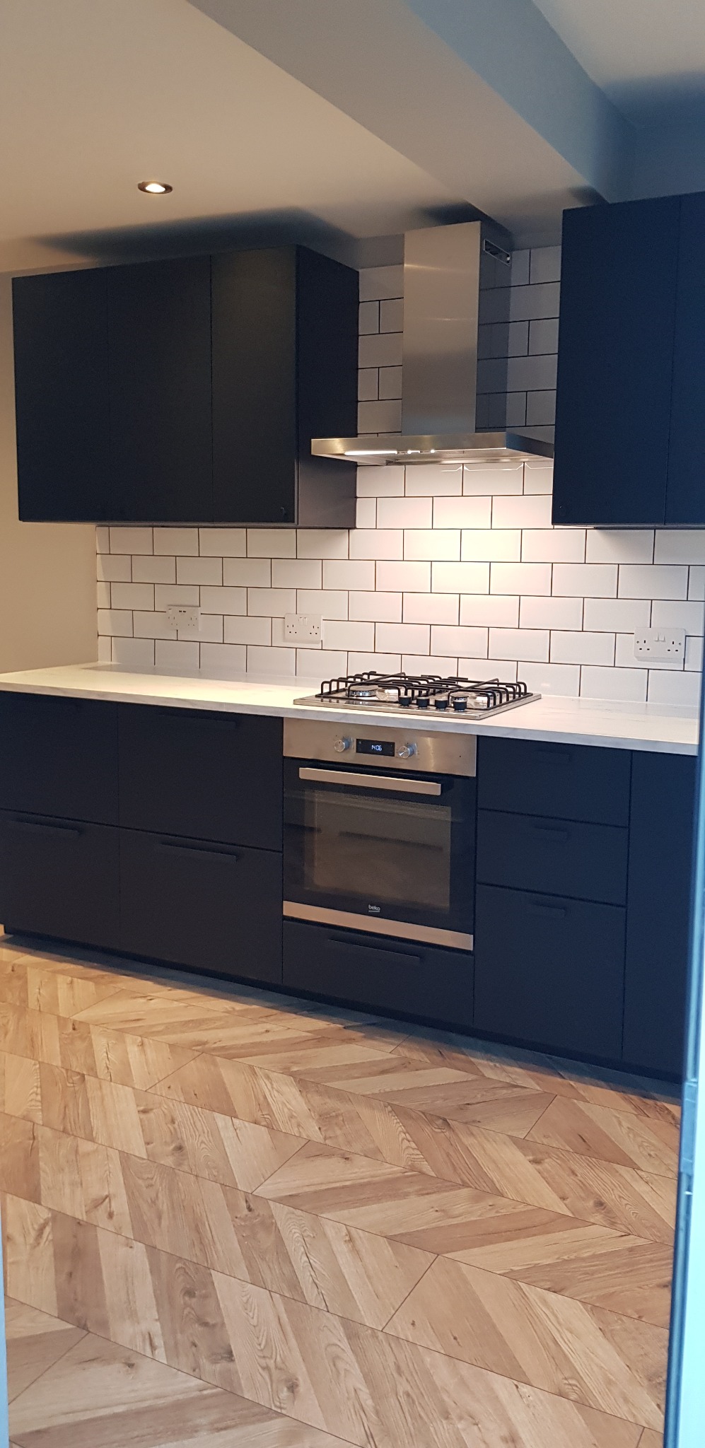 Ciemna zabudowa kuchenna z białym blatem i płytkami typu metro, wykończona stalowym okapem i piekarnikiem, na podłodze parkiet w jodełkę.