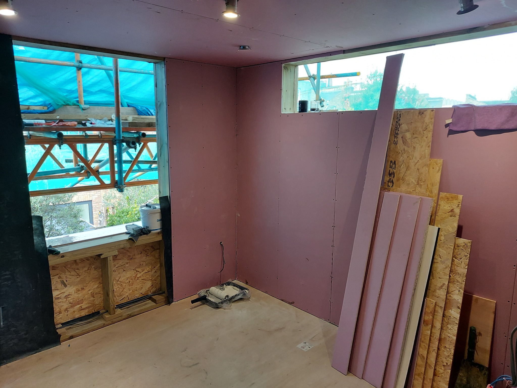 Wnętrze w trakcie prac wykończeniowych: ściany pokryte różowymi płytami, widoczne profile, okno z rusztowaniem na zewnątrz, narzędzia malarskie na podłodze, przygotowane płyty OSB.