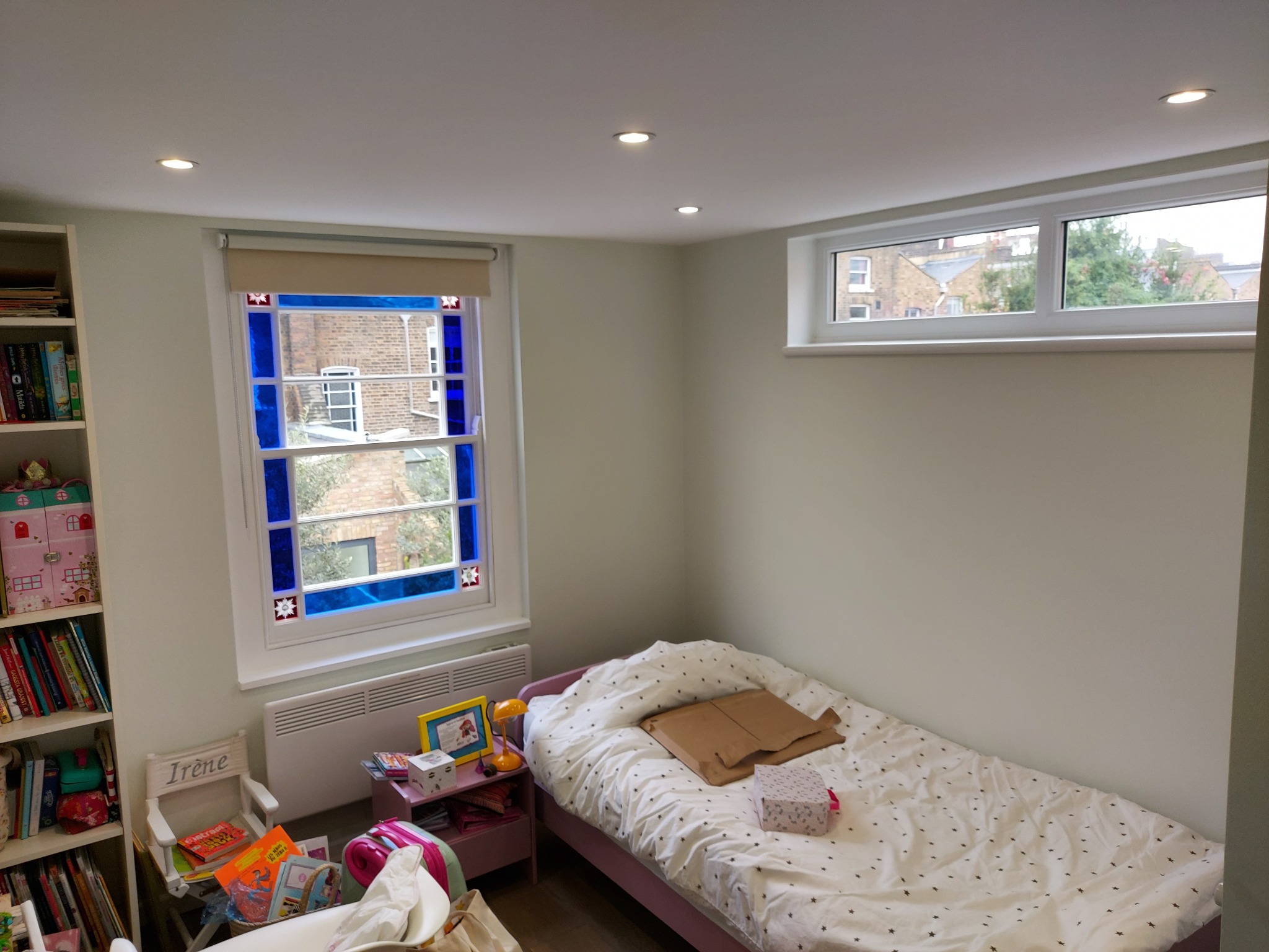 Pokój dziecięcy z łóżkiem z motywem gwiazdek, kolorowym oknem z niebieskimi akcentami i wbudowanym oświetleniem punktowym w suficie.