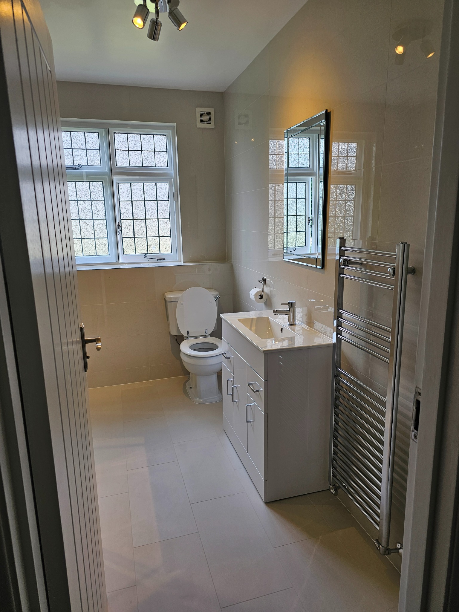 Wykończona łazienka w Bielsku-Białej z beżowymi płytkami, białym zestawem toaletowym, szafką pod umywalką i grzejnikiem drabinkowym, widoczna zza uchylonych drzwi.