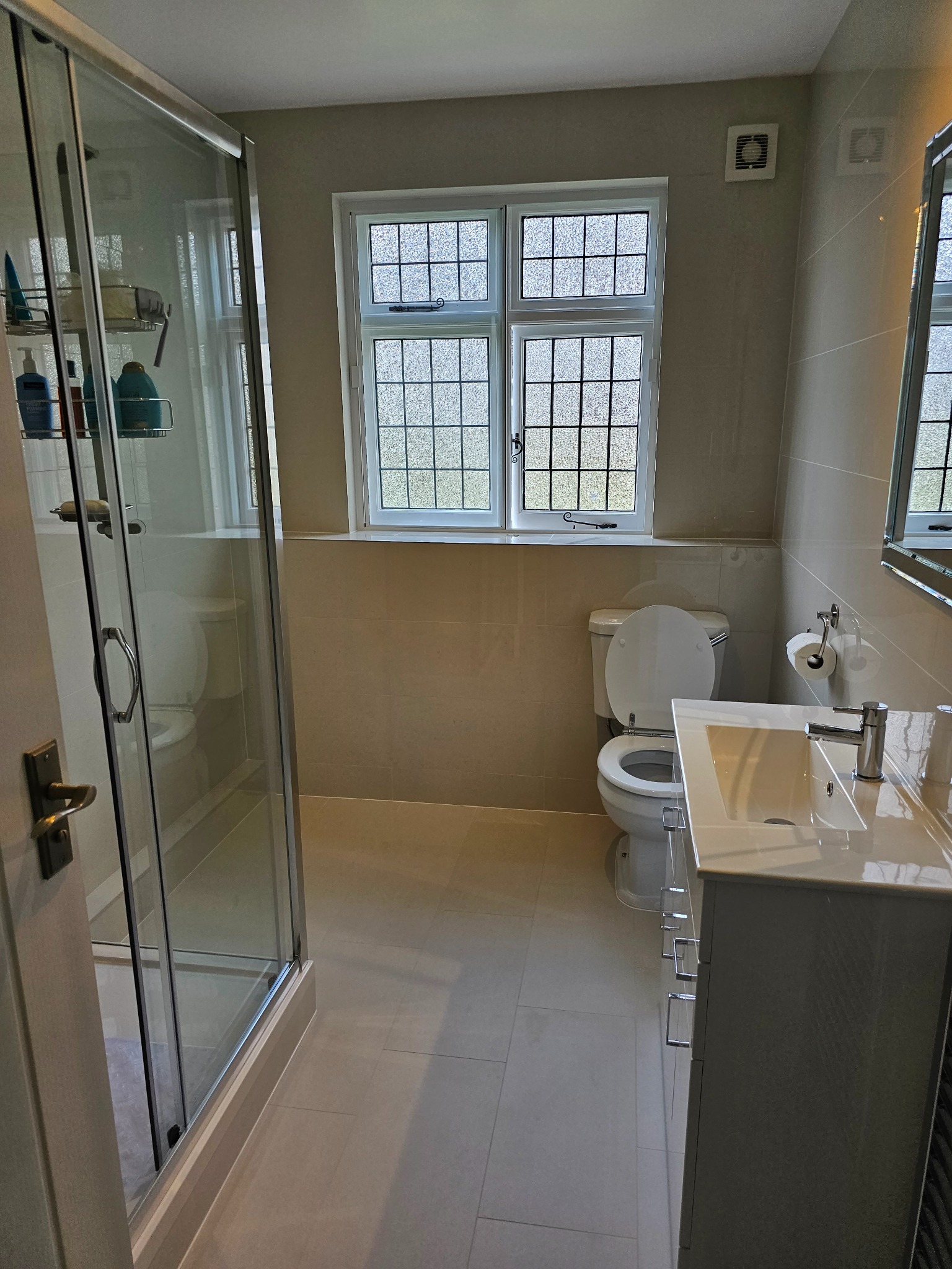 Jasna łazienka z kabiną prysznicową, umywalką z szafką z szufladami, toaletą i oknem z ozdobnymi szybami, wykończona beżowymi płytkami.