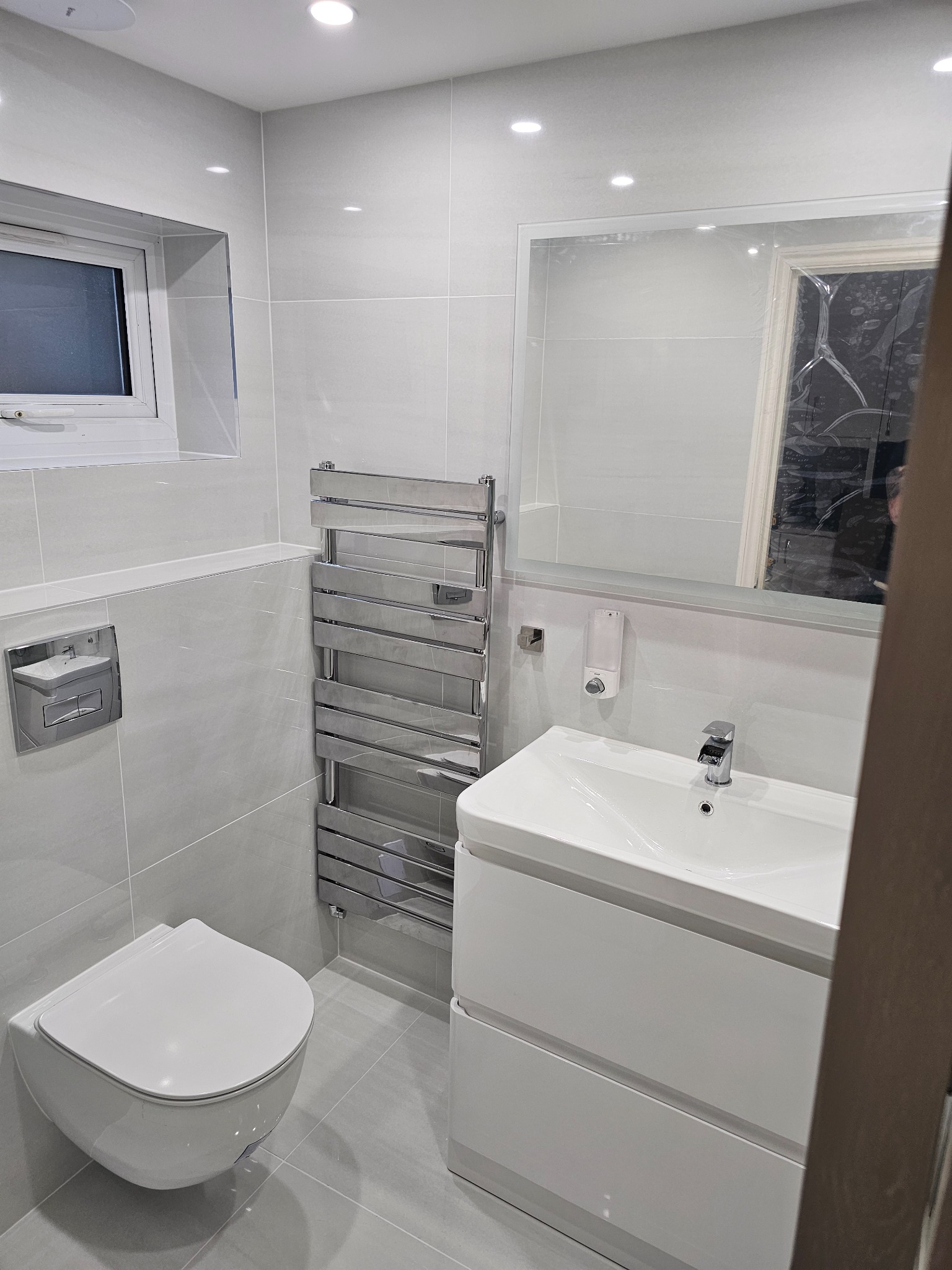 Nowoczesna łazienka w odcieniach szarości z podwieszaną miską WC, grzejnikiem drabinkowym i szafką umywalkową z dwiema szufladami, widok zza futryny drzwi.