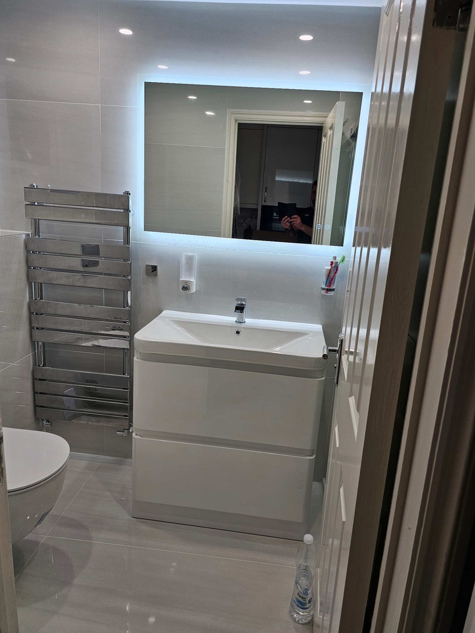 Nowoczesna łazienka w odcieniach szarości z podświetlanym lustrem nad białą szafką umywalkową, grzejnik drabinkowy po lewej stronie, widok zza uchylonych drzwi.