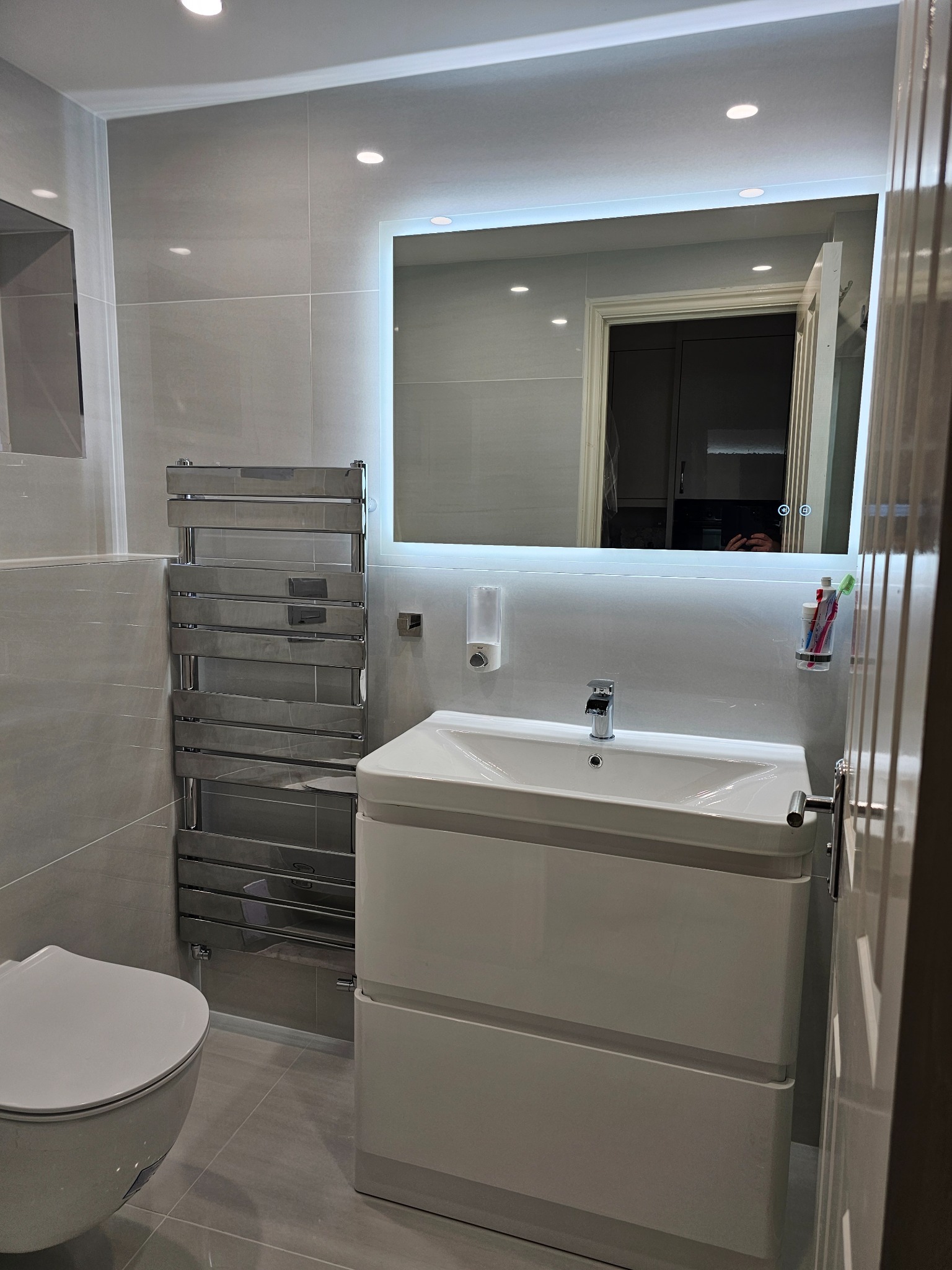 Wykończona łazienka w odcieniach szarości z podświetlanym lustrem, grzejnikiem drabinkowym i białą szafką pod umywalką, widok na fragment toalety i drzwi.