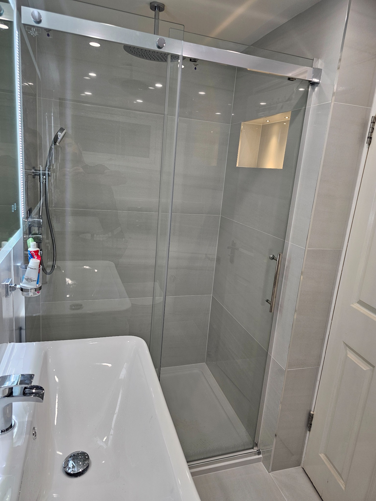 Nowoczesna łazienka z szarą płytką, kabiną prysznicową ze szklanymi drzwiami przesuwnymi, widoczna umywalka i armatura, oświetlenie halogenowe w suficie i wnęce prysznicowej.