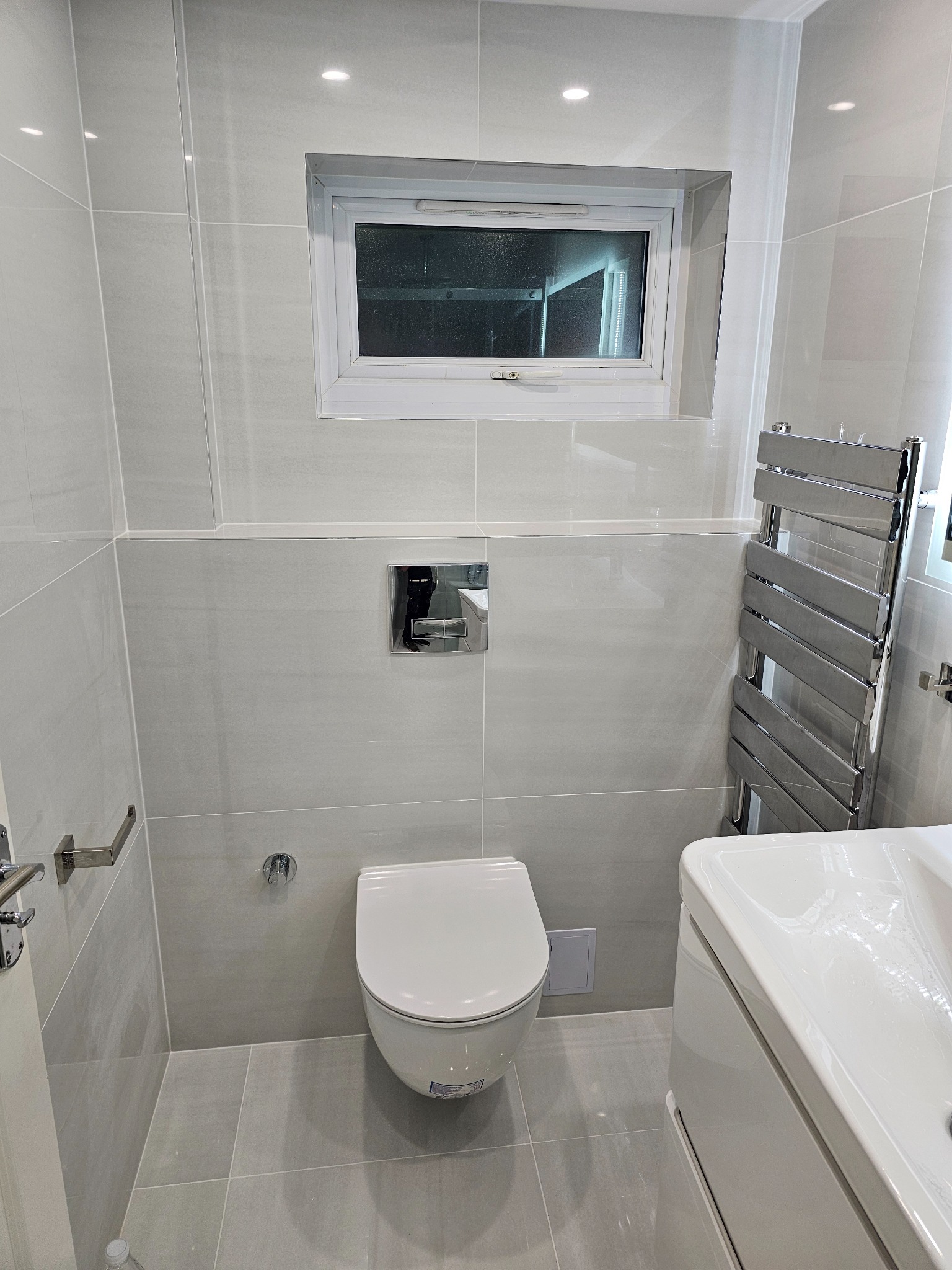 Nowoczesna, jasna łazienka po remoncie, z szarymi płytkami, podwieszaną toaletą, grzejnikiem drabinkowym i oknem.