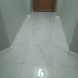 Błyszczące, białe płytki podłogowe z szarym marmurkowym wzorem, ułożone w korytarzu z drewnianymi drzwiami i szarozielonymi ścianami, wykończone białymi listwami przypodłogowymi.