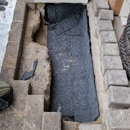 Do Usług Andrzej Ziółkowski Usługi Ogólnobudowlane - Przygotowanie podłoża pod kostkę brukową z warstwą hydroizolacji przy ścianie budynku, widoczne fragmenty kostki, piasek i narzędzia.