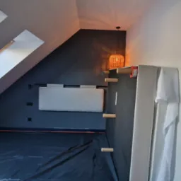 Poddasze z oknem dachowym zabezpieczone folią malarską, widoczna ściana pomalowana na granatowo z zamontowanym panelem tapicerowanym i lampą z wikliny, w tle widoczny fragment pracownika.