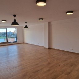 Do Usług Andrzej Ziółkowski Usługi Ogólnobudowlane - Jasne, przestronne wnętrze z drewnianą podłogą, dużym oknem i nowoczesnym oświetleniem sufitowym; osoba widoczna w przejściu do innego pomieszczenia.