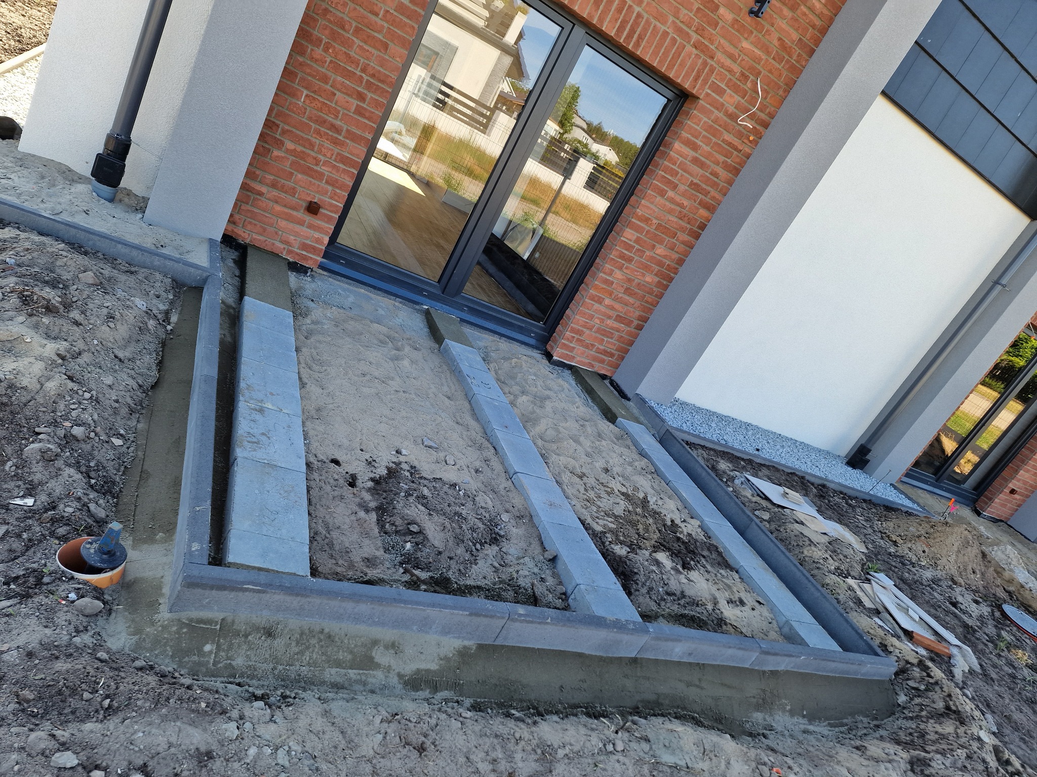 Przygotowanie podłoża pod taras z obrzeżami z kostki betonowej, widoczne piasek i ziemia, w tle elewacja budynku z cegły i okno.