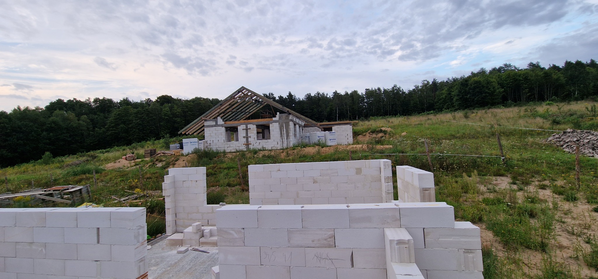 Budowa domu jednorodzinnego w stanie surowym otwartym, widoczne ściany z bloczków, więźba dachowa, teren zalesiony w tle.