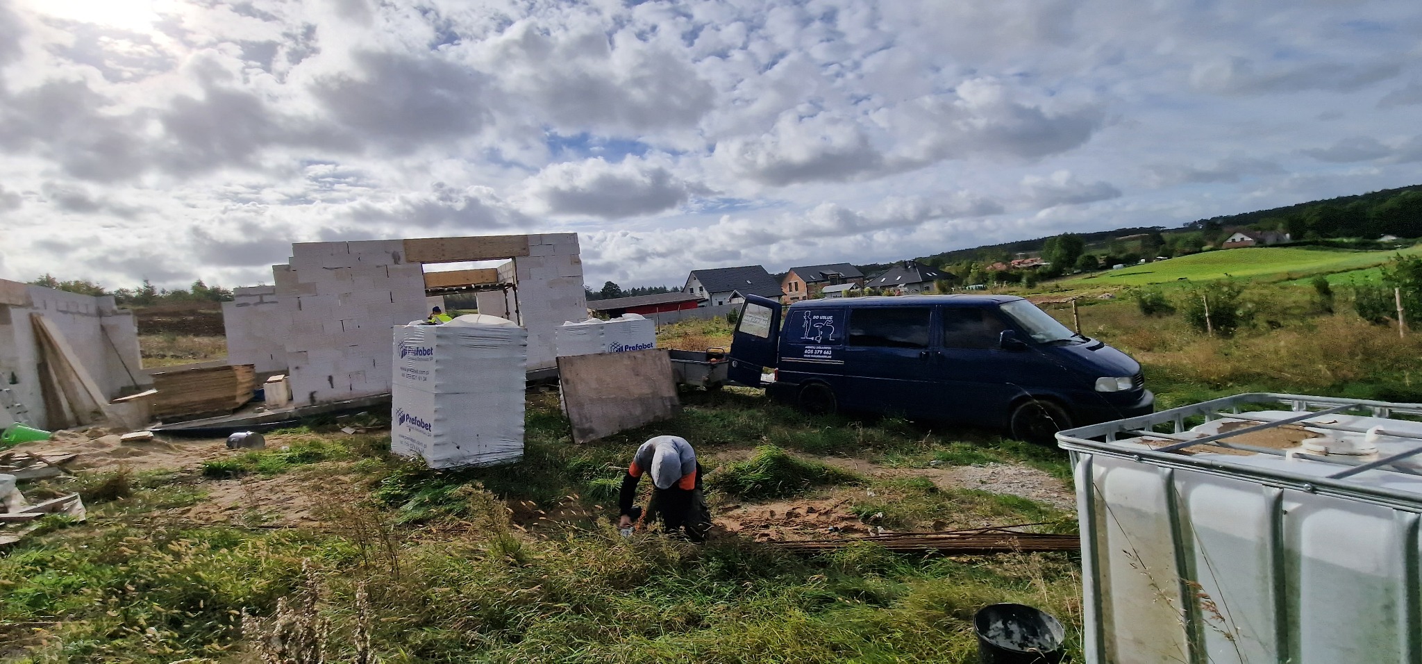 Budowa domu jednorodzinnego w trakcie wznoszenia ścian z bloczków, widoczny pracownik, samochód dostawczy z otwartymi drzwiami i materiały budowlane na działce.
