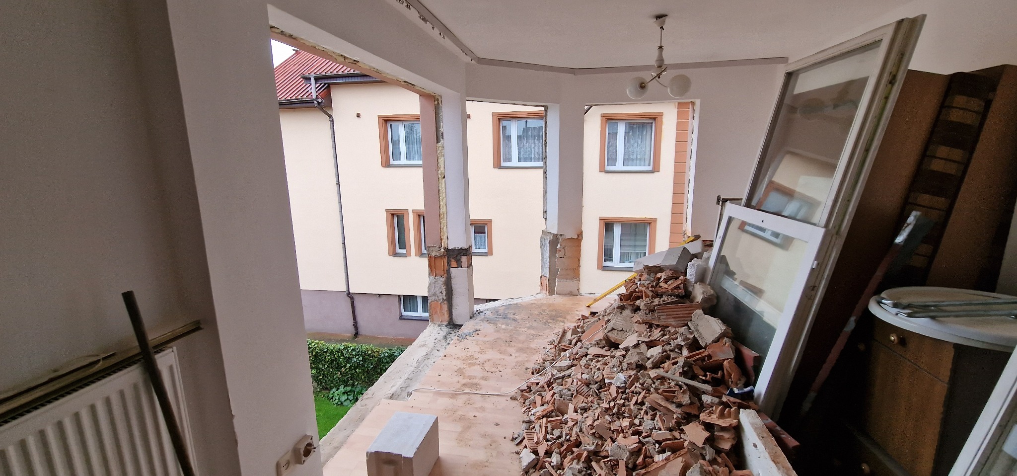 Demontaż balkonu: sterta gruzu ceglanego na balkonie z widokiem na budynek mieszkalny, stare okna oparte o ścianę.