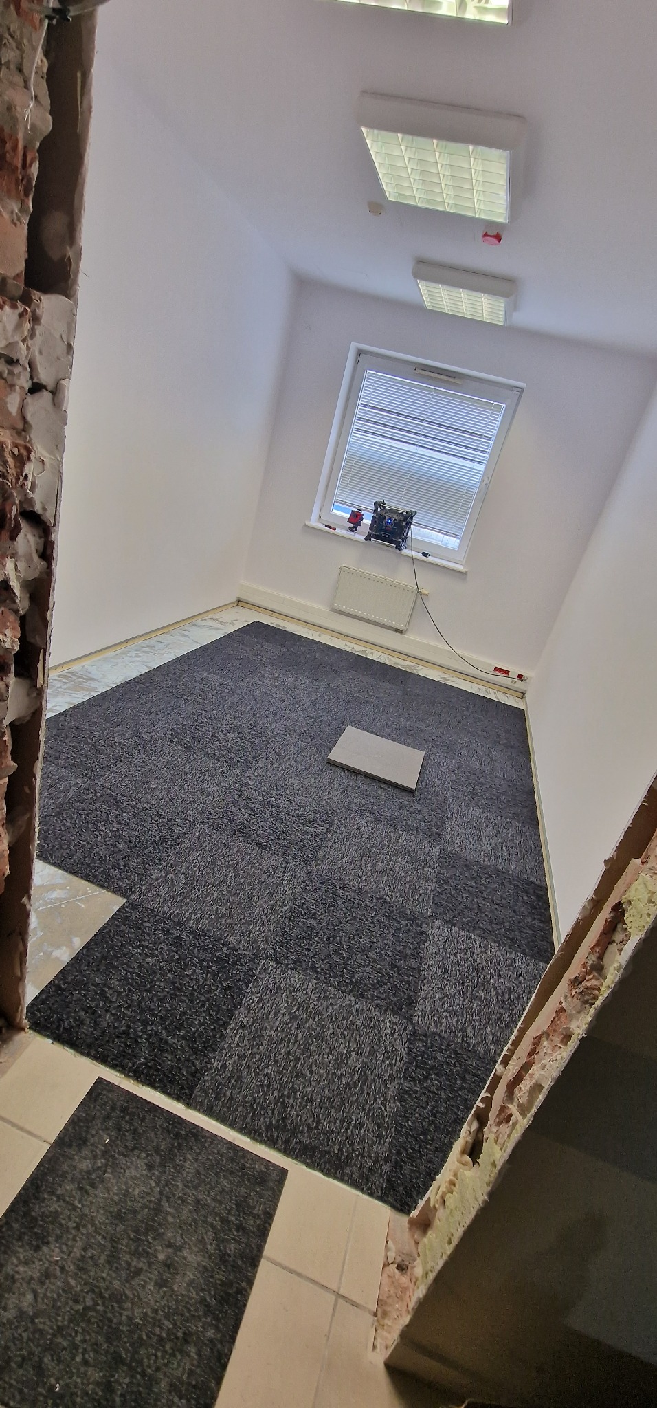 Układanie ciemnoszarej wykładziny dywanowej w kwadratach w pomieszczeniu biurowym z oknem dachowym i widocznymi fragmentami surowych ścian.