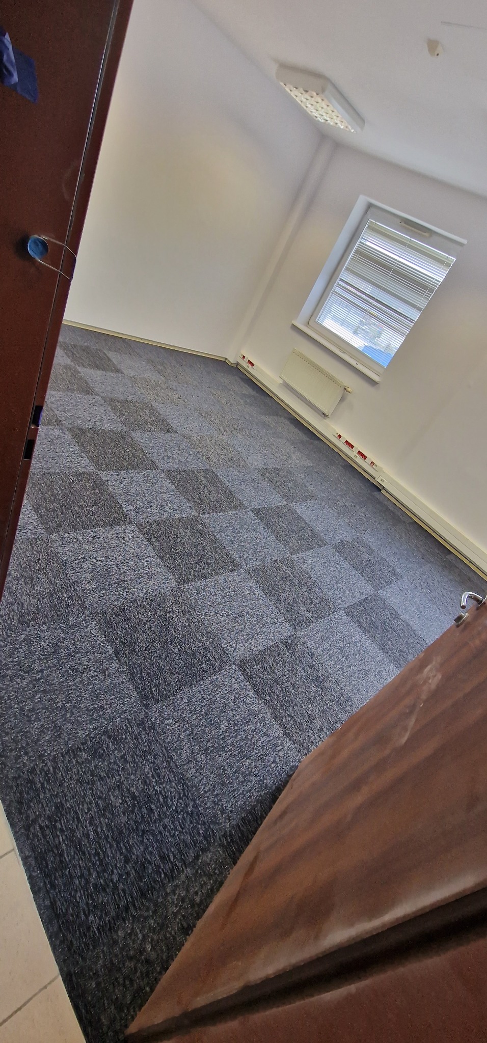 Wnętrze biura z nową, szaro-czarną wykładziną dywanową w kwadraty, widok zza otwartych, drewnianych drzwi.