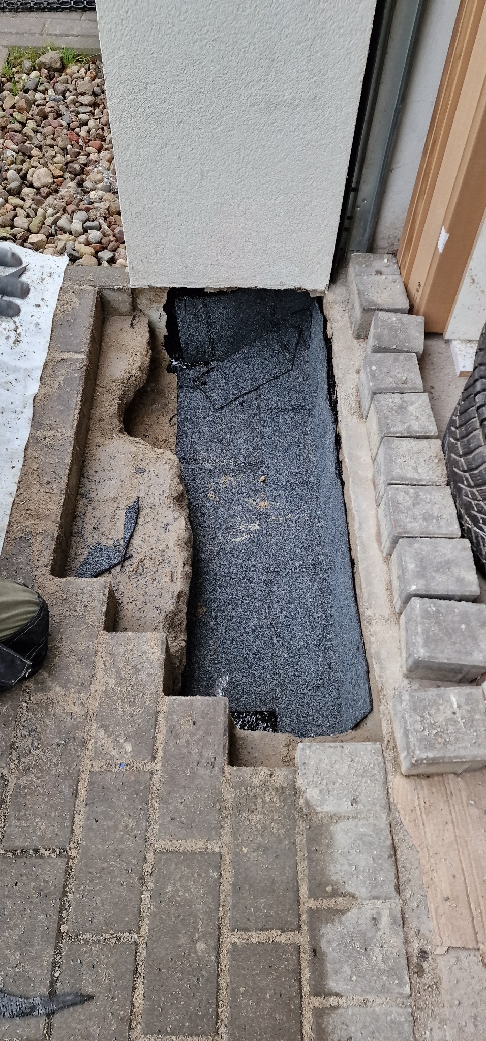 Przygotowanie podłoża pod kostkę brukową z warstwą hydroizolacji przy ścianie budynku, widoczne fragmenty kostki, piasek i narzędzia.