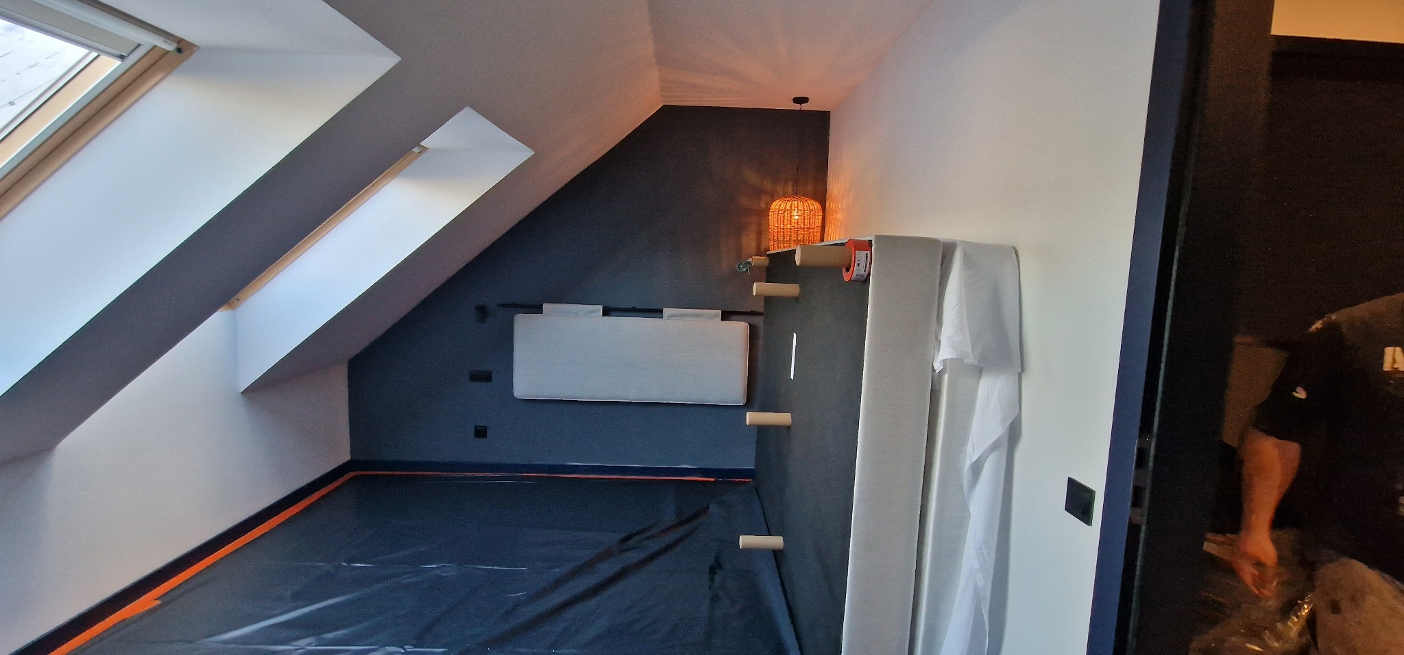 Poddasze z oknem dachowym zabezpieczone folią malarską, widoczna ściana pomalowana na granatowo z zamontowanym panelem tapicerowanym i lampą z wikliny, w tle widoczny fragment pracownika.