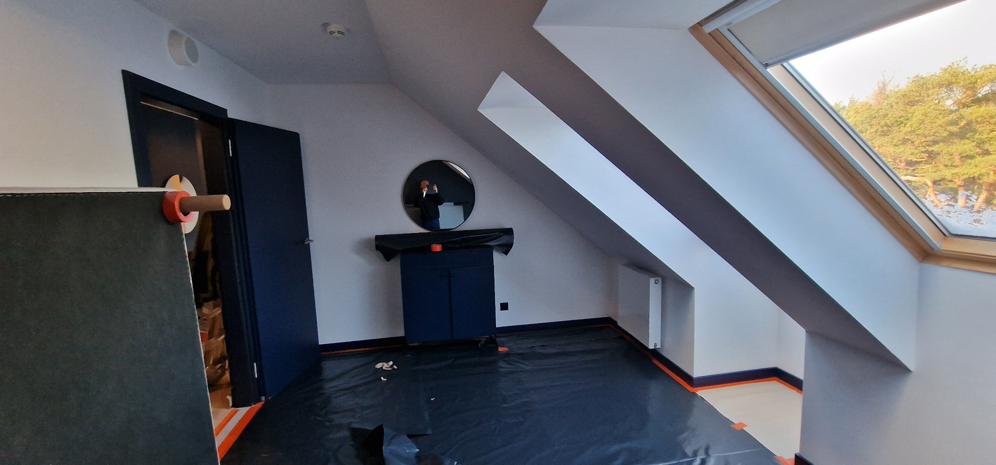 Pokój na poddaszu w trakcie remontu: zabezpieczona podłoga folią, widoczne okno dachowe, drzwi i szafka z lustrem.