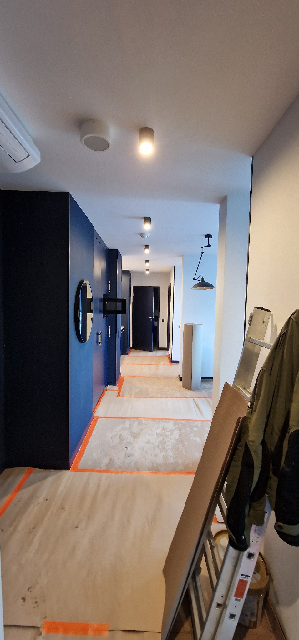 Korytarz w trakcie remontu z zabezpieczoną podłogą, widoczne zabudowane szafy w kolorze granatowym, lustro, lampy sufitowe, drabina i wisząca lampa.