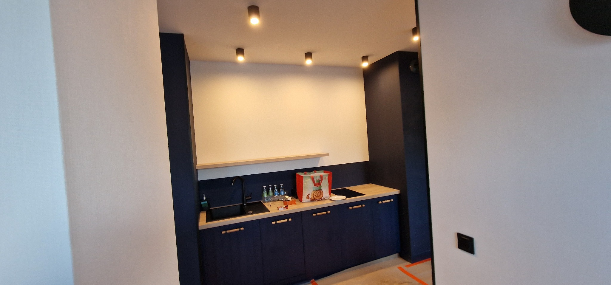 Nowoczesna, granatowa zabudowa kuchenna z drewnianym blatem i półką, oświetlona punktowymi lampami sufitowymi, widoczna z perspektywy wejścia do pomieszczenia.