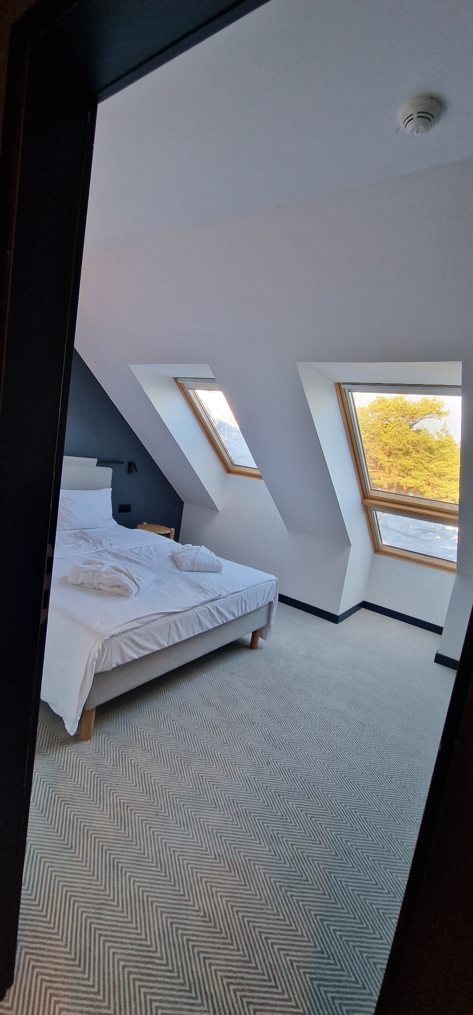 Pokój na poddaszu z dwoma oknami dachowymi, łóżkiem z białą pościelą i dywanem w zygzaki, widok zza framugi drzwi.