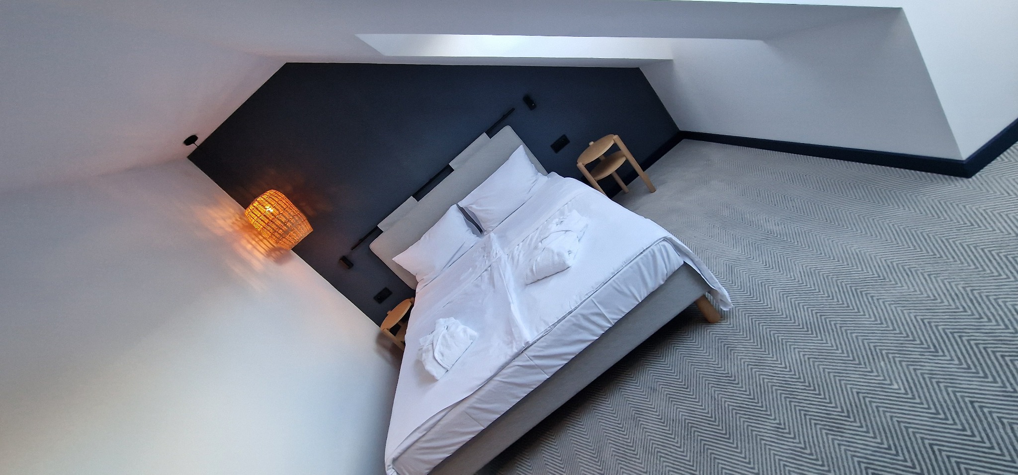 Pokój na poddaszu z łóżkiem, lampką nocną z wikliny i dywanem w jodełkę.