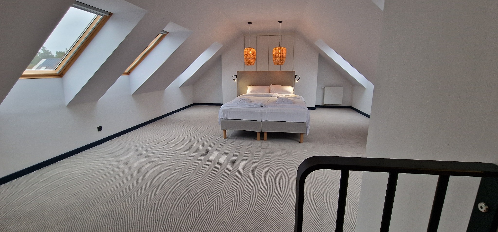Jasne poddasze z łóżkiem, oknami dachowymi i dywanem w jodełkę, widok zza czarnej balustrady.