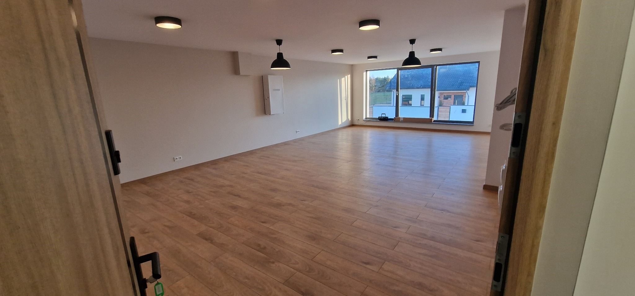 Puste, jasne pomieszczenie z drewnianą podłogą, widok zza otwartych drzwi, nowoczesne oświetlenie sufitowe, duże okna z widokiem na dom.
