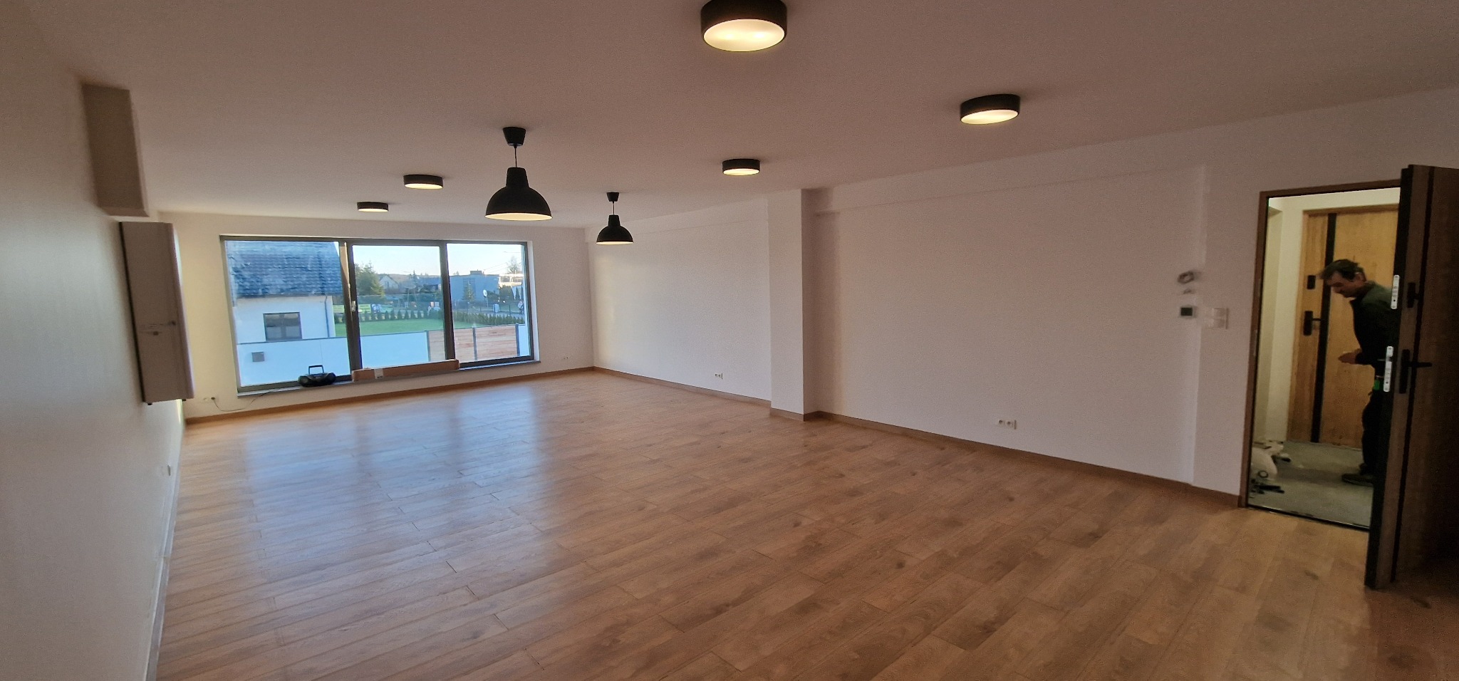 Jasne, przestronne wnętrze z drewnianą podłogą, dużym oknem i nowoczesnym oświetleniem sufitowym; osoba widoczna w przejściu do innego pomieszczenia.