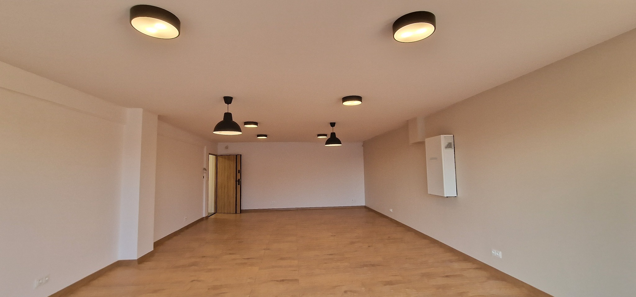 Wnętrze nowo wykończonego pokoju z drewnianą podłogą, białymi ścianami i nowoczesnym oświetleniem sufitowym, w tym czarne lampy wiszące i okrągłe lampy wpuszczane.