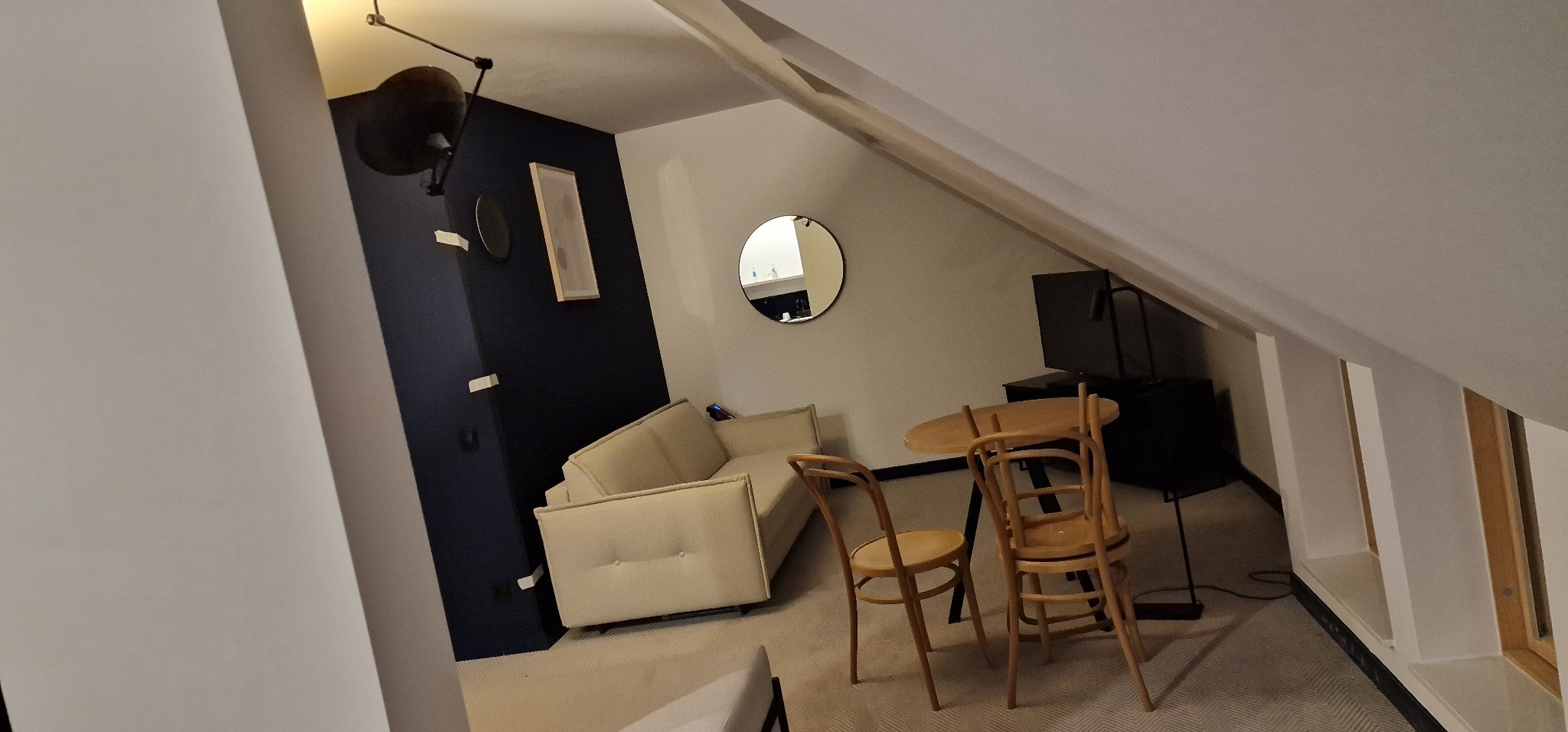 Przestrzeń pod skosem z kremową sofą, okrągłym lustrem, drewnianym stołem i krzesłami, oświetlona lampą na ścianie w kolorze granatowym.
