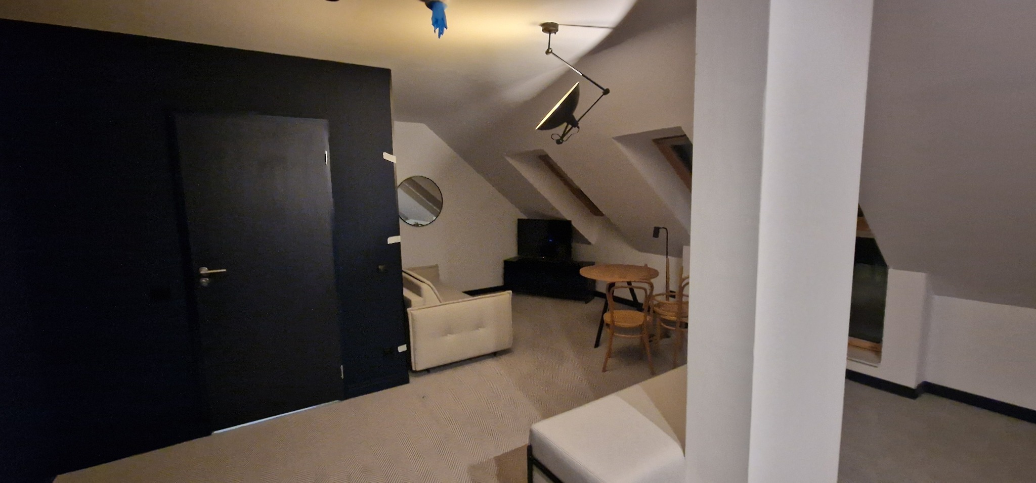 Ukończone poddasze w stylu minimalistycznym z czarnymi drzwiami, okrągłym lustrem, jasną sofą, drewnianym stolikiem i krzesłami, oświetlone nowoczesną lampą sufitową.