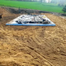 Przygotowany teren pod wylewkę fundamentów, widoczna stal zbrojeniowa pod czarną folią, szalunek z niebieskiego styroduru, taczka z narzędziami w tle, ziemia z wyraźnymi śladami opon.