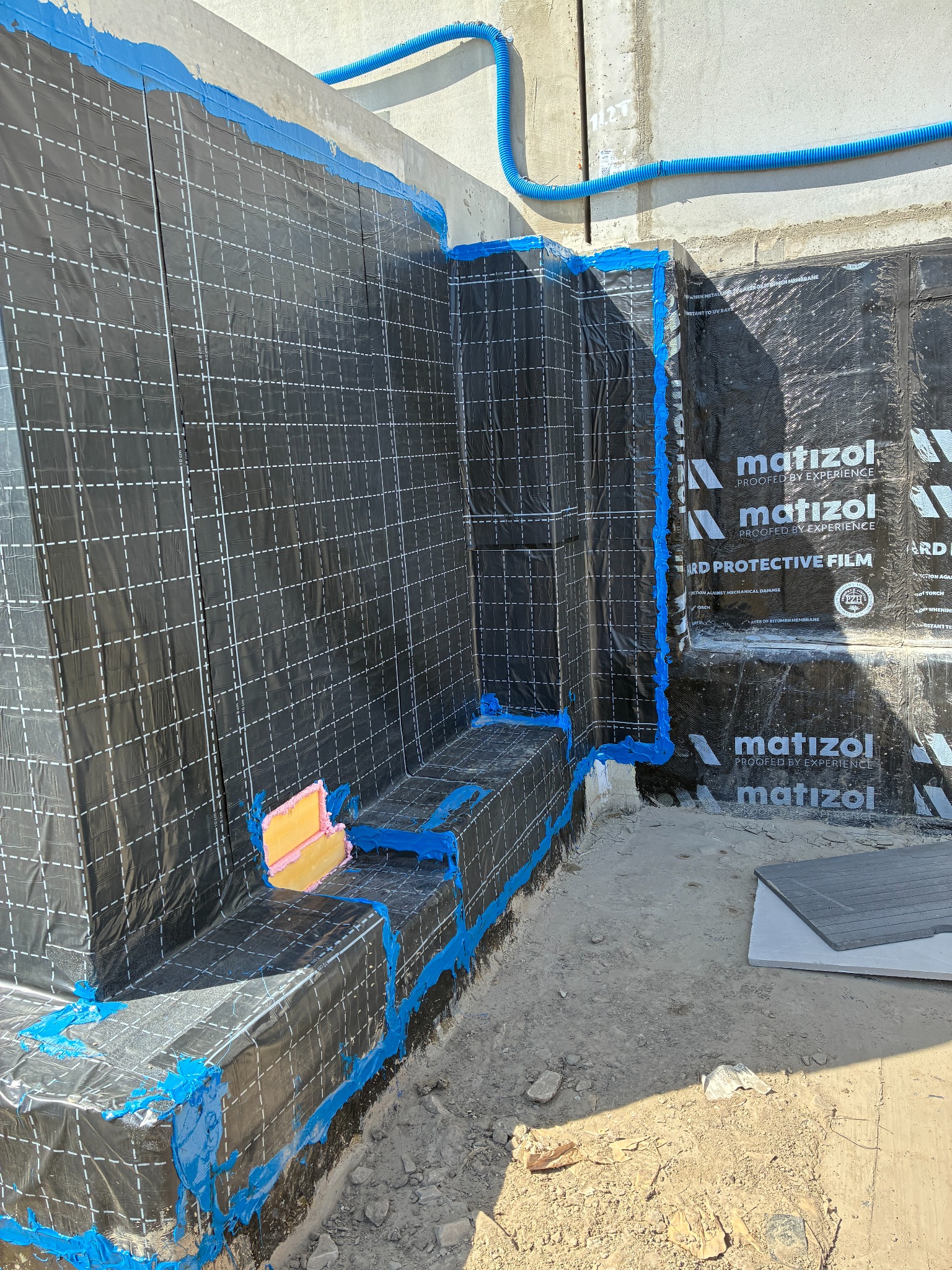Izolacja fundamentów z folią Matizol i niebieską taśmą uszczelniającą. Widoczna rura drenażowa. Prace izolacyjne w toku, z detalami mocowania i zabezpieczeń.