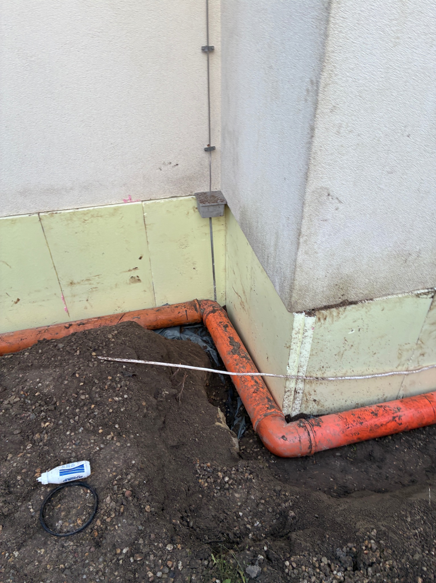 Pomarańczowe rury drenażowe ułożone w wykopie przy fundamencie budynku z izolacją. Widoczny grunt, uszczelniacz i czarna folia. Ujęcie z bliska, z dołu.