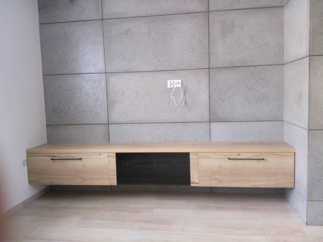 Wisząca szafka RTV z drewnianymi frontami i czarną wnęką, zamontowana na ścianie wyłożonej szarymi panelami betonowymi z widocznym gniazdkiem elektrycznym.