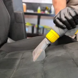 Czyszczenie ciemnej, welurowej tapicerki samochodowej za pomocą odkurzacza ekstrakcyjnego z żółtą końcówką, widoczne czarne rękawice ochronne.