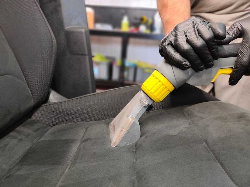 Czyszczenie ciemnej, welurowej tapicerki samochodowej za pomocą odkurzacza ekstrakcyjnego z żółtą końcówką, widoczne czarne rękawice ochronne.