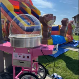 Różowy wózek do waty cukrowej marki King na trawie, z dmuchańcem Wild West w tle, podczas imprezy plenerowej w słoneczny dzień. Dziecięce obuwie obok wózka.