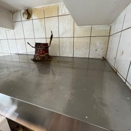 Myjnia parowa Legnica Wojciech Jurkiewicz - Zanieczyszczona powierzchnia robocza ze stali nierdzewnej w pomieszczeniu z brudnymi, białymi płytkami ceramicznymi na ścianach. Widoczny resztki po czyszczeniu lub zalaniu, oraz wiadro...