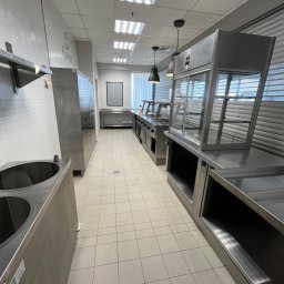 Myjnia parowa Legnica Wojciech Jurkiewicz - Wnętrze profesjonalnej kuchni z nierdzewnymi urządzeniami do serwowania posiłków, widok w głąb pomieszczenia z oświetleniem sufitowym i jasnymi kafelkami na podłodze.
