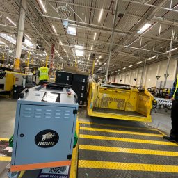 Myjnia parowa Legnica Wojciech Jurkiewicz - Wnętrze hali magazynowej w Legnicy z wózkiem transportowym Fortador i żółtym wózkiem widłowym, widoczne oznakowanie poziome i pracownik w odblaskowej kamizelce.