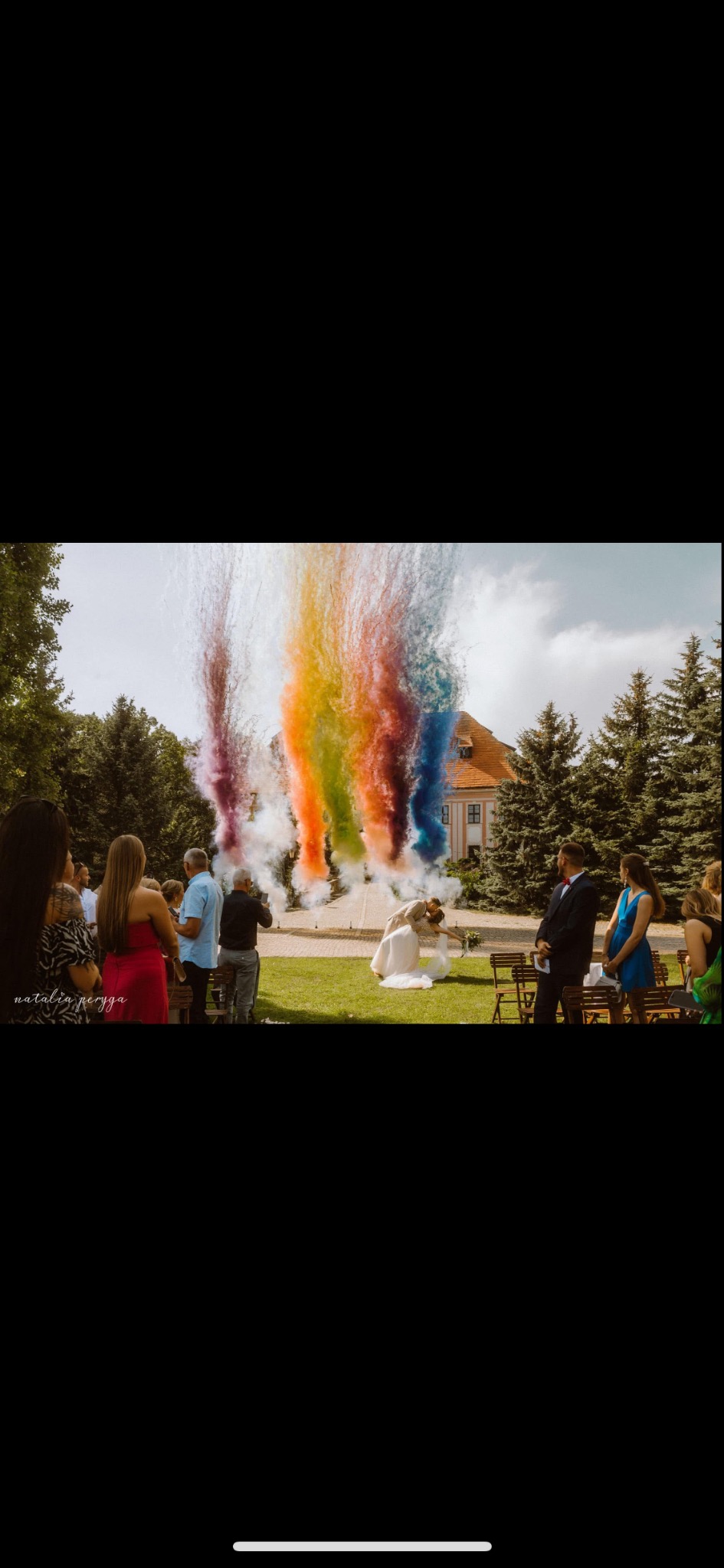 Para młoda klęczy na trawie wśród gości, a za nimi tęczowe fontanny dymu podczas ceremonii ślubnej w plenerze z budynkiem w tle.
