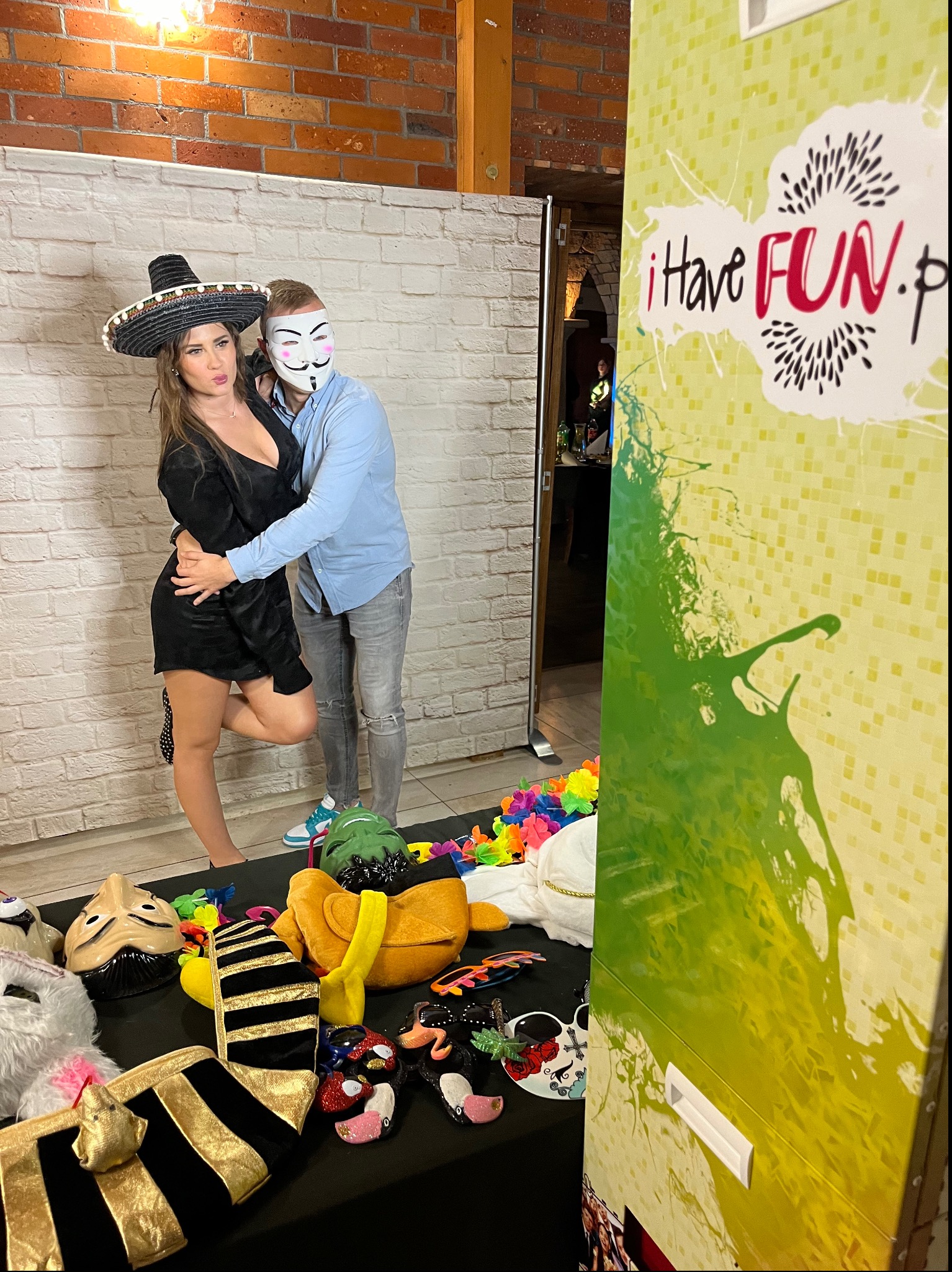 Para pozuje w fotobudce z rekwizytami: kobieta w sombrero i mężczyzna w masce. Na stole leżą różne maski i akcesoria. W tle ceglana ściana i grafika fotobudki.