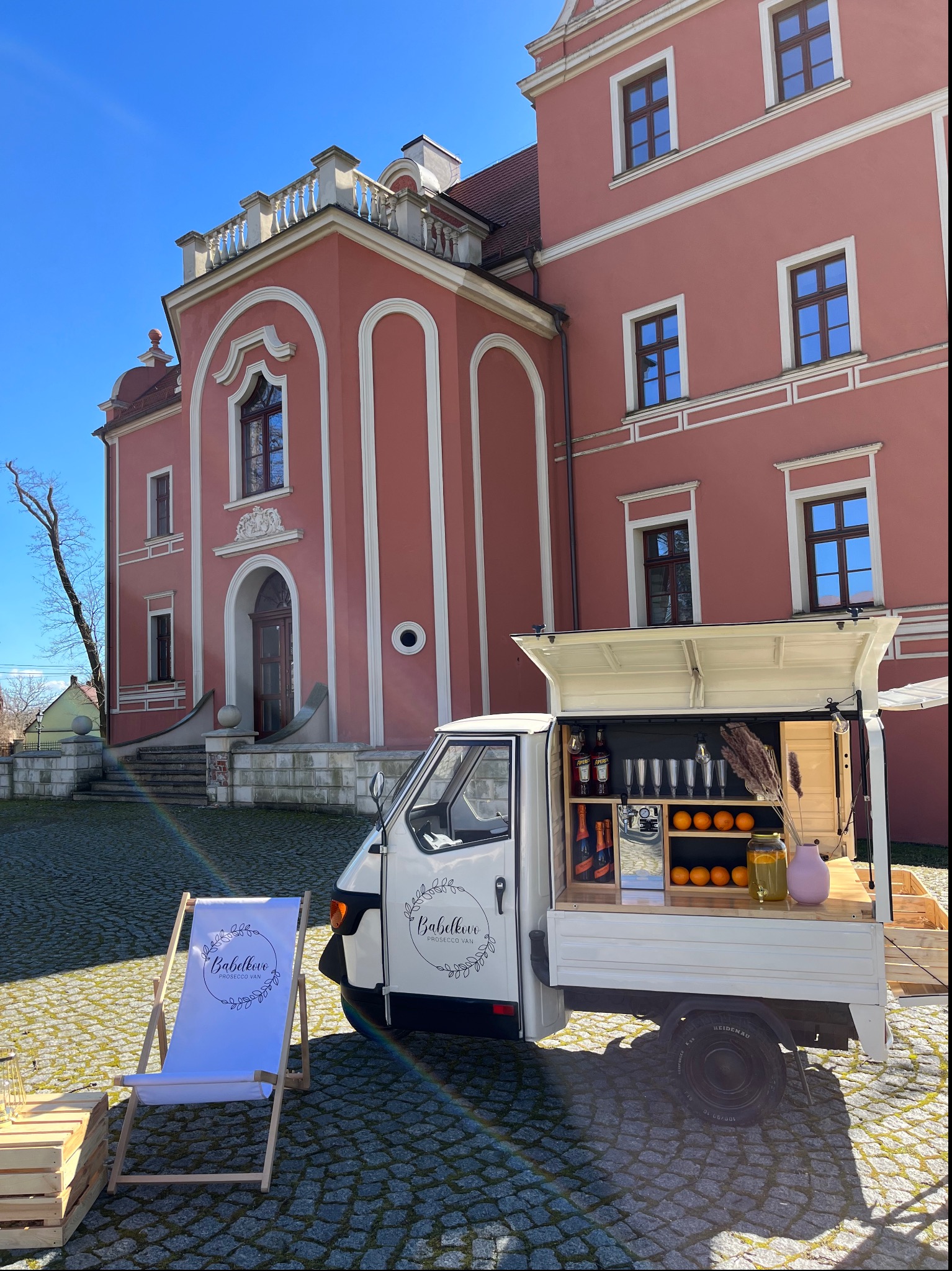 Mobilny bar Prosecco Van na tle różowego budynku z białymi zdobieniami, uzupełniony leżakiem z logo. Słoneczny dzień, kostka brukowa.