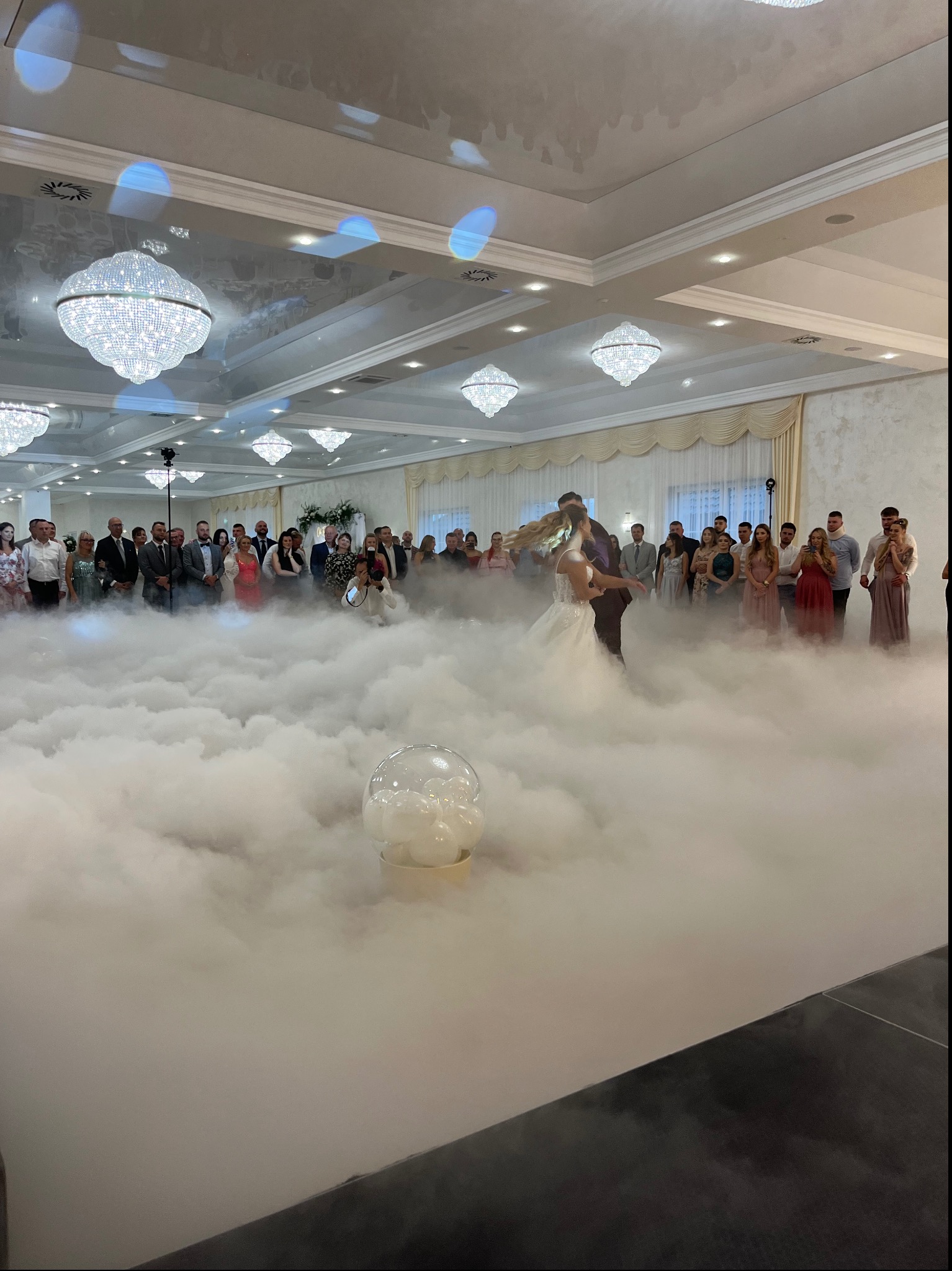 Pierwszy taniec pary młodej w dymie na weselu, goście w tle, kryształowe żyrandole. Romantyczna atmosfera, dekoracje sali weselnej.