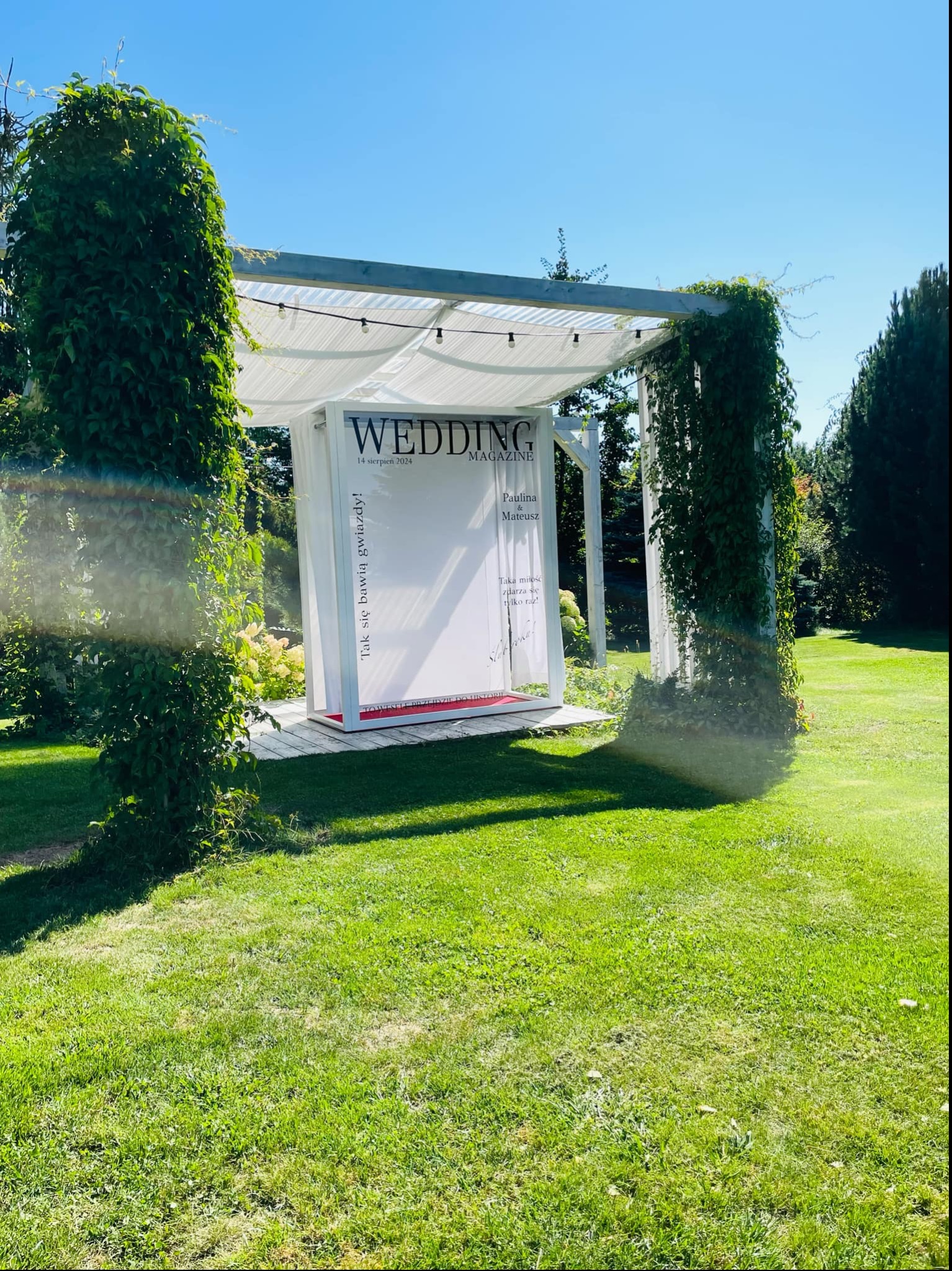Plenerowa instalacja ślubna: biała altana z napisem 'Wedding Magazine', opleciona zielenią, na tle trawnika i drzew. Dekoracja na imprezę.