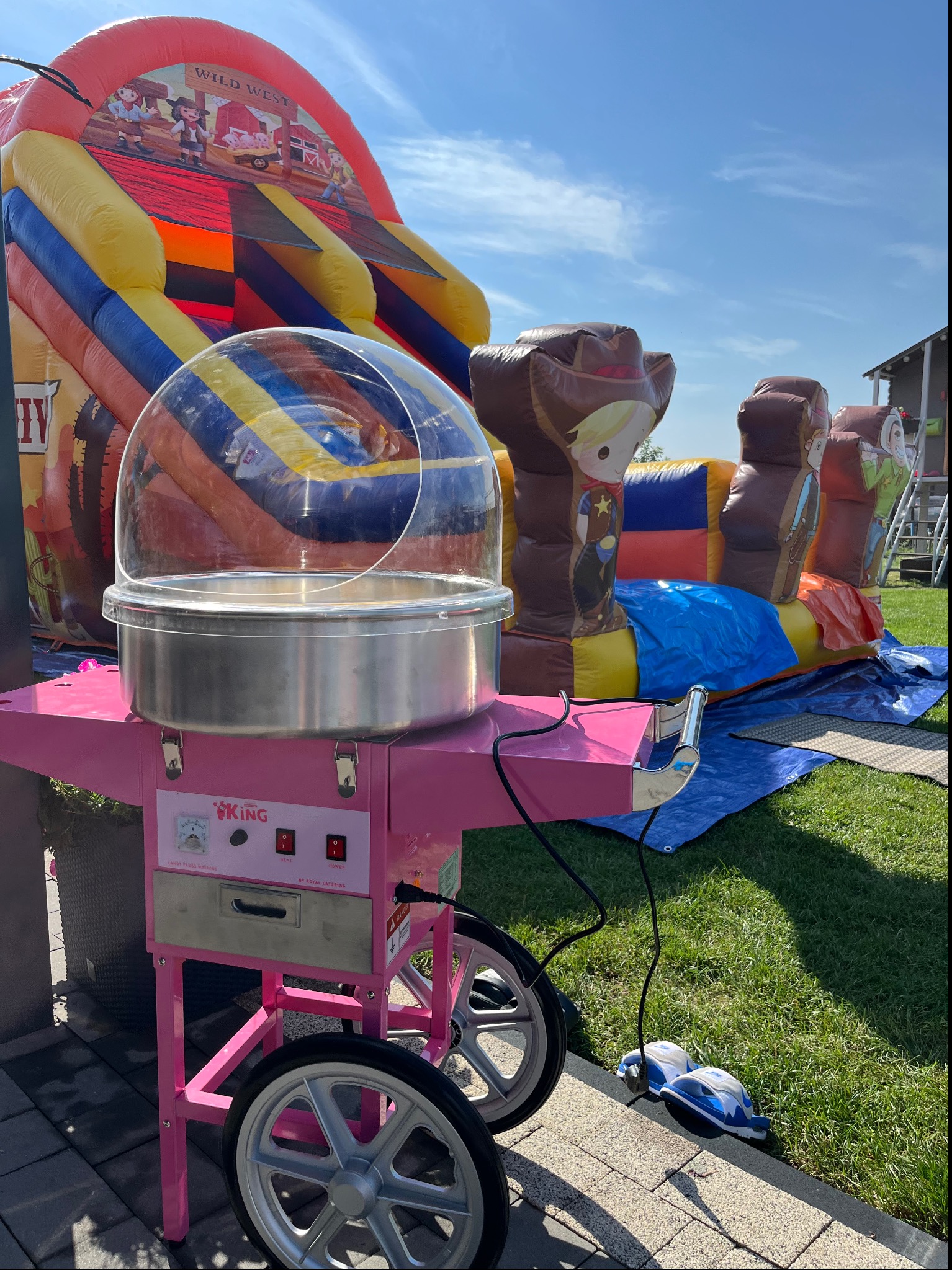 Różowy wózek do waty cukrowej marki King na trawie, z dmuchańcem Wild West w tle, podczas imprezy plenerowej w słoneczny dzień. Dziecięce obuwie obok wózka.