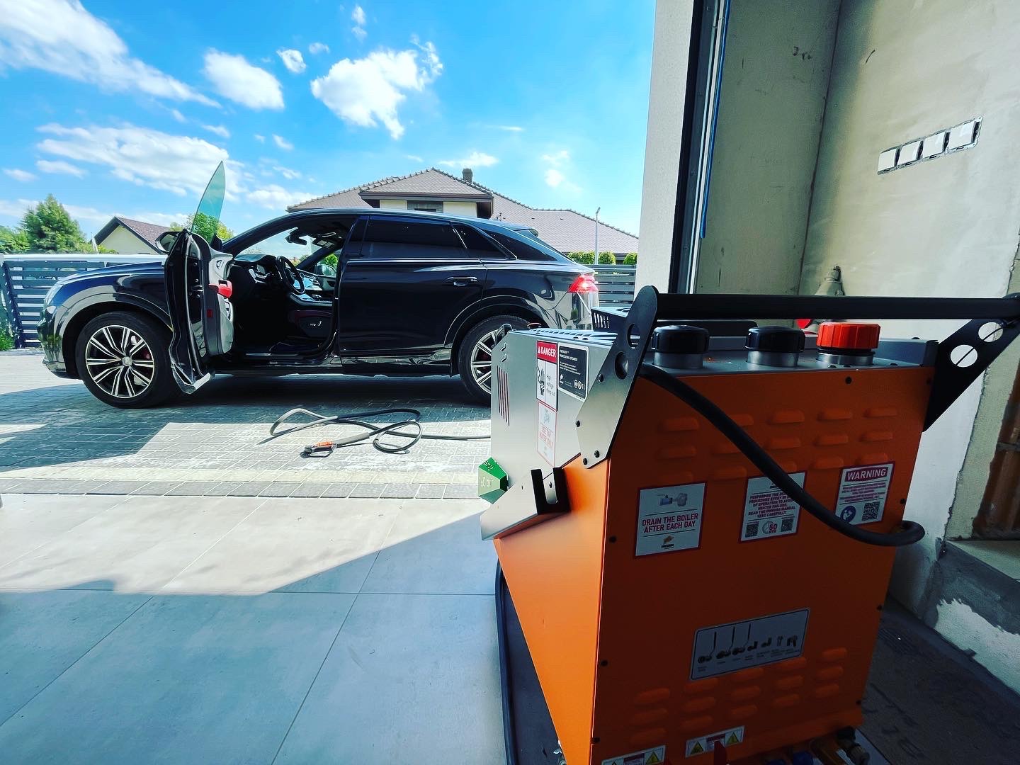 Czarny SUV z otwartymi drzwiami kierowcy, zaparkowany na jasnoszarej kostce brukowej przed garażem; obok stoi pomarańczowe urządzenie czyszczące z naklejkami ostrzegawczymi i wężem.